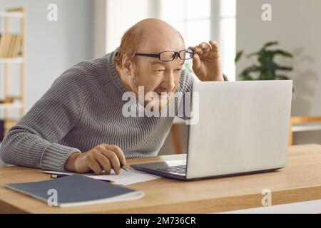 Uomo maturo confuso che ha una visione scarsa e lavora sul notebook a casa indossando gli occhiali. Foto Stock