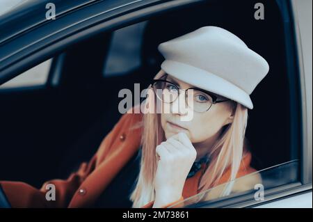 Un'elegante donna di mezza età che indossa un cappello grigio e occhiali si siede dietro il volante della sua auto Foto Stock