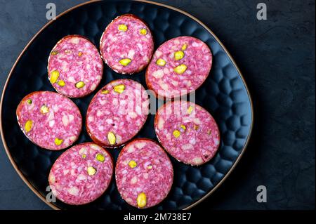 Salame tradizionale affumicato con spezie e pistacchi al nero. Salame fette di salsiccia su un piatto nero. Sfondo scuro. Foto Stock
