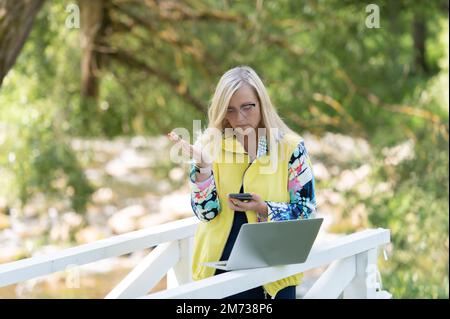 Donna in viaggio d'affari con notebook e telefono cellulare che comunicano in remoto nel parco cittadino Foto Stock