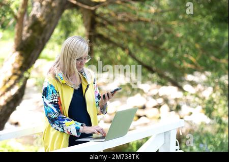 Attraente donna di mezza età che utilizza smartphone e computer portatile all'aperto Foto Stock