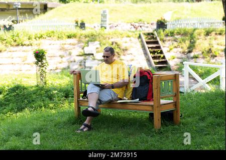 Uomo di mezza età seduto sulla panca di legno nel parco e utilizzando un computer portatile, lavorando all'aperto Foto Stock