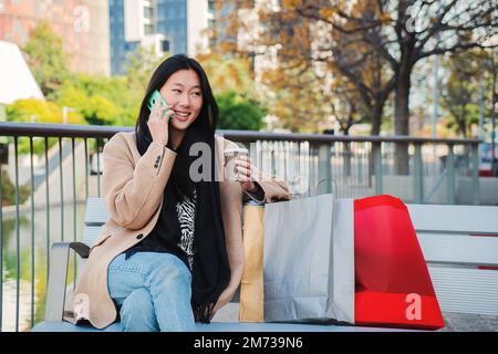 Felice ragazza asiatica che chiama utilizzando un telefono cellulare seduto su una panca. Una donna cinese che sorride e parla con lo smartphone con le borse della spesa all'aperto. Concetto di comunicazione. Foto di alta qualità Foto Stock