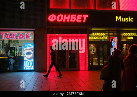 Un giovane che cammina lungo il marciapiede passa accanto alle finestre dei negozi illuminate di notte. Via Novy Arbat (New Arbat), Mosca, Russia. Foto Stock