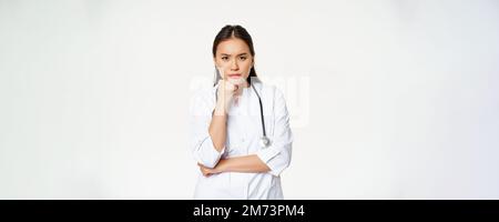 Immagine di infermiera arrabbiata, medico asiatico femminile che guarda infastidito e infastidito, sopracciglia solco e solcante pensieroso, in piedi su sfondo bianco Foto Stock