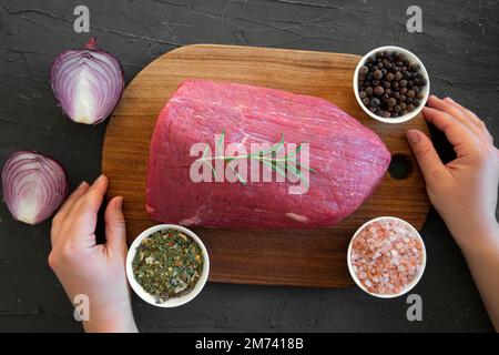 Carne rossa cruda su sfondo testurizzato in marmo nero, vista dall'alto. Il tagliere tiene le mani femminili. Foto Stock