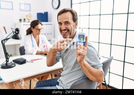 Uomo di mezza età in clinica medico con dispositivo glucometro sorridente felice di puntare con la mano e il dito Foto Stock