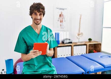 Giovane uomo ispanico che indossa uniforme fisioterapista utilizzando il touchpad presso la clinica fisioterapista Foto Stock
