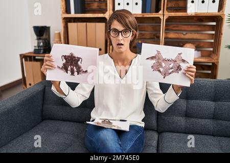 Bruna donna che lavora con rorschach test in psicologia clinica in shock faccia, guardando scettico e sarcastico, sorpreso con la bocca aperta Foto Stock