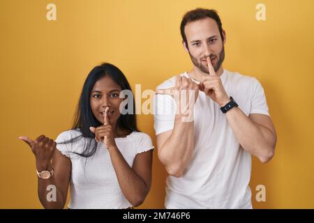 Coppia interrazziale in piedi su sfondo giallo chiedendo di essere tranquilla con il dito sulle labbra che punta con la mano a lato. silenzio e concetto segreto. Foto Stock