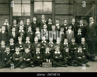 Londra. circa. 1912. Una classe di giovani scolari e il loro insegnante posano per una fotografia di gruppo davanti al loro edificio scolastico. I ragazzi frequentano la Gopsall Street School di Hackney e sono orgogliosi di indossare le medaglie di frequenza della London School, che includono la ‘Medaglia del Re Edoardo VII’ e il suo successore, la ‘Medaglia del Re’. Un ragazzo sostiene una tavola con il nome della classe, “Classe i”. La rete metallica copre le finestre per evitare danni da giochi di palla. Molti dei ragazzi indossano rigidi collari di lino. Foto Stock