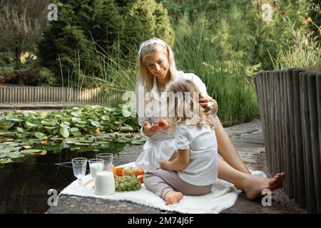 Donna gentile e attraente che nutra la bambina al picnic, con frutta fresca biologica e pranzo al latte nel parco verde vicino al lago Foto Stock