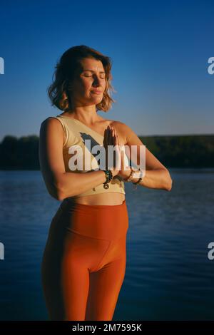 Una donna yoga sta facendo il sole saluto vicino all'acqua su un molo al tramonto. Foto Stock