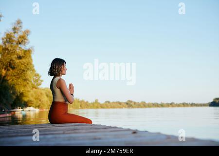 La donna Yogi pratica esercizi di respirazione sul molo mentre tiene le mani in una posa namaste. Foto Stock