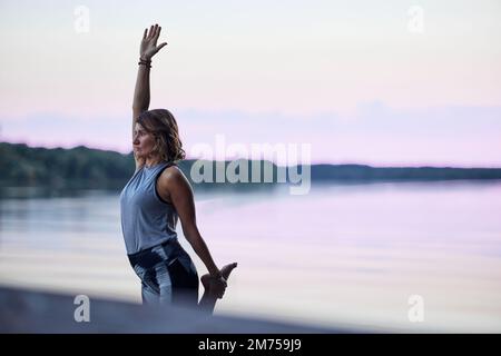 Una donna pratica lo yoga e l'equilibrio su un piede sul molo vicino al fiume in natura. Foto Stock