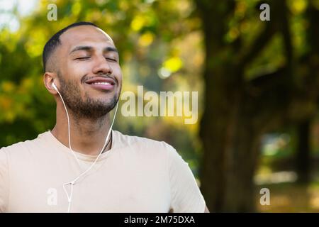 Primo piano ritratto di sportivo nel parco, uomo ispanico jogging nel parco con gli occhi chiusi respirando aria fresca e riposando, jogging con cuffie ascoltando musica, radio online e podcast. Foto Stock