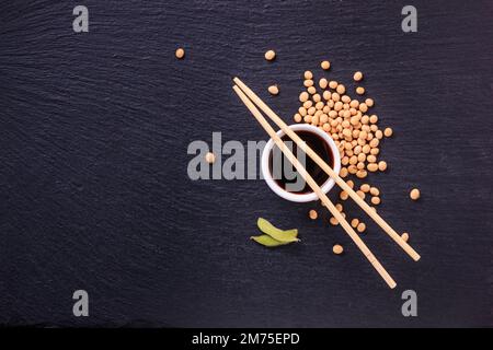 Salsa di soia in una ciotola con bastoncini di bambù e semi di soia sparsi sulla superficie nera della pietra ardesia, vista dall'alto, primo piano con spazio per il testo Foto Stock