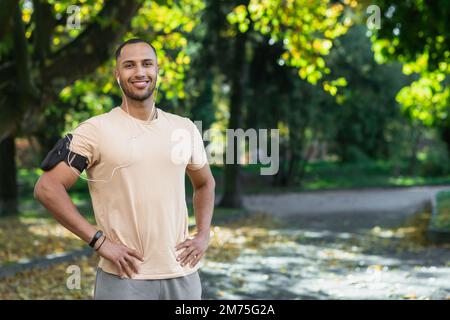 Ritratto dell'uomo di successo nel parco uomo ispanico con cuffie che ascoltano musica e audiolibri podcast online, sorridendo e guardando la macchina fotografica mentre jogging e fare esercizio fisico. Foto Stock