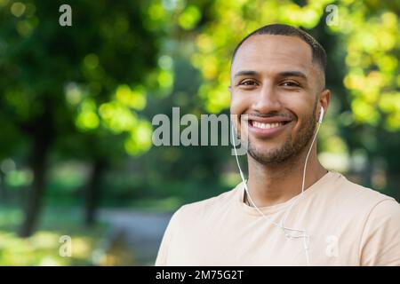 Ritratto dell'uomo di successo nel parco uomo ispanico con cuffie che ascoltano musica e audiolibri podcast online, sorridendo e guardando la macchina fotografica mentre jogging e fare esercizio fisico. Foto Stock