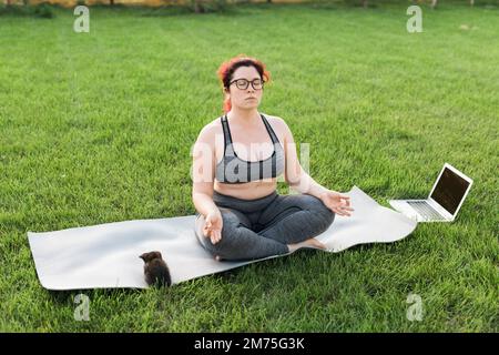 Più misura positività del corpo donna che fa meditazione su tappetino yoga con gattino sul cortile - concetto di benessere e salute mentale Foto Stock