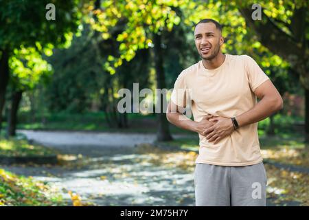 Atleta latino-americana ha un grave dolore allo stomaco, l'uomo che tiene la mano a fianco dello stomaco dopo aver fatto esercizio fisico e fitness nel parco esterno. Foto Stock