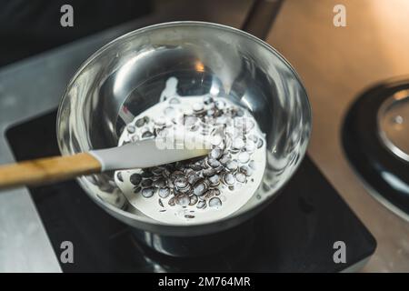 Sciogliere le gocce di cioccolato. Gocce di cioccolato fondente sciolte con panna densa in ciotola di acciaio inossidabile con una spatola. Scatto in interni. Foto di alta qualità Foto Stock