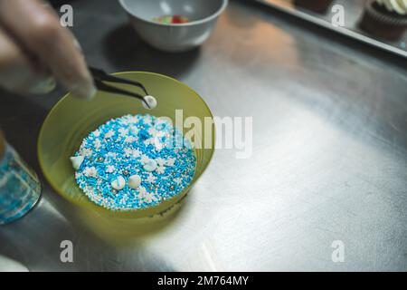 Fornaio professionale che usa le pinzette per decorare i pasticcini. Processo di decorazione dei cupcake. Guarnizioni bianche e blu. Primo piano. Foto di alta qualità Foto Stock