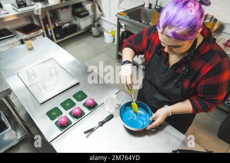 Workshop sulla decorazione della torta. Giovane donna che prepara glassa blu o crema di burro glassa in una ciotola di acciaio. Scatto da interno ad angolo alto. Spazio di copia. Foto di alta qualità Foto Stock