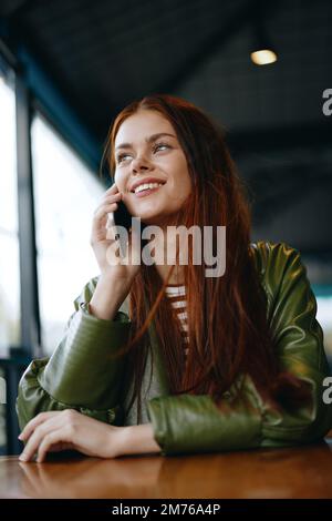 Donna con i capelli rossi blogger freelance seduto in un caffè che parla al telefono e sorridente, ragazza hipster in stile di vita di abiti alla moda Foto Stock