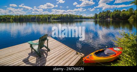 Sedia seduta su una banchina di legno di fronte ad un lago calmo con una canoa rossa in estate Foto Stock