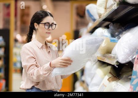 Ritratto di donna caucasica con occhiali sceglie cuscino e biancheria da letto. Tessili per la casa in supermercato. Concetto di shopping. Foto Stock