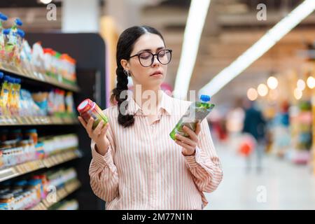 Ritratto di giovane donna caucasica che indossa occhiali tiene i prodotti in mano e non può fare scelta. Concetto di shopping in supermercato e consumismo. Foto Stock