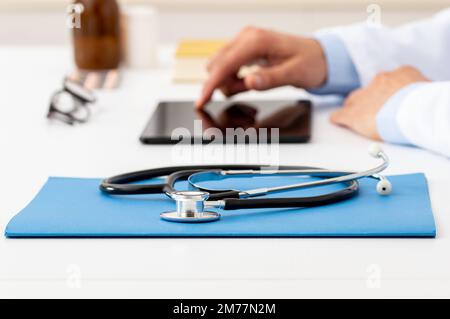 Primo piano di uno stetoscopio appoggiato su un tablet con un medico irriconoscibile che lavora in background con un computer portatile Foto Stock