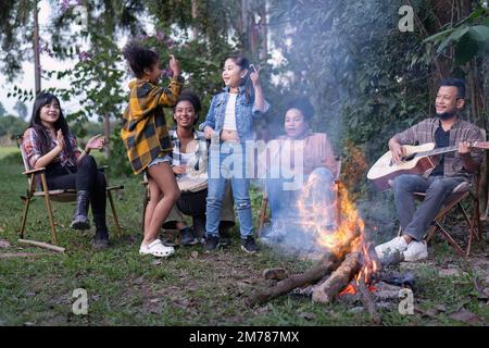 Gruppo di camper di famiglia seduti accanto a salsicce grigliate al fuoco e danze cantanti si divertono Foto Stock