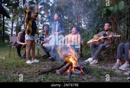 Gruppo di camper di famiglia seduti accanto a salsicce grigliate al fuoco e danze cantanti si divertono Foto Stock