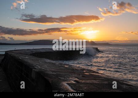 L'onda si schiantava sulla fine del Cobb Dorset Inghilterra con un'alba arancione brillante sul mare sullo sfondo Foto Stock