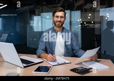 Ritratto di un giovane bell'uomo, freelance, uomo d'affari, ragioniere. Seduto in ufficio, guardando la fotocamera, sorridendo. Funziona su un computer portatile, con documenti, compila i moduli con una penna. Foto Stock