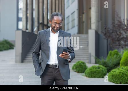 Un bell'uomo d'affari afro-americano è in piedi vicino a un centro uffici, tenendo un telefono nelle sue mani. Digitare messaggi, effettuare chiamate, attendere una riunione. Tiene la mano in tasca, sorride. Foto Stock