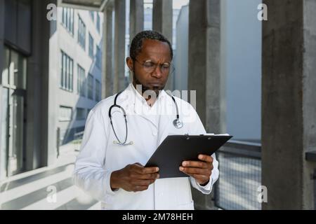 Un medico afro-americano preoccupato e penoso sta in piedi vicino all'ospedale, tenendo una cartella nelle sue mani. Egli legge ansiosamente le carte, la storia medica del paziente. Foto Stock