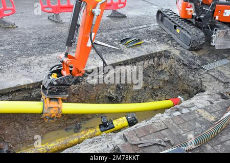 Morsetto idraulico mini-scavatore montato sul braccio posteriore dell'attore che afferra 125mm tubo principale del gas di plastica giallo di ricambio spinto e tirato nel vecchio tubo di acciaio UK Foto Stock