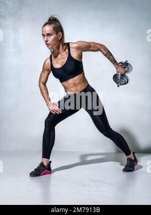 Donna sportiva che si allena con i manubri. Foto del modello in abbigliamento sportivo nero su sfondo grigio. Motivazione sportiva e stile di vita sano Foto Stock