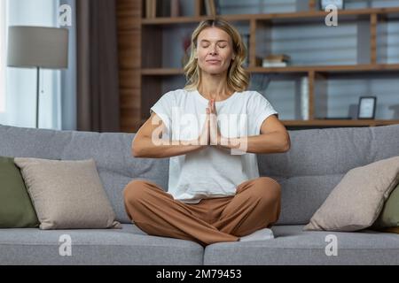 Donna anziana a casa riposo meditando con gli occhi chiusi seduta sul divano in soggiorno, casalinga bionda matura in loto posa sorridente. Foto Stock