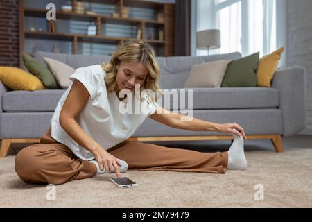 Donna anziana attiva sorridente sul pavimento che fa allungamenti ed esercizi, utilizzando il telefono e l'app di allenamento online a casa nel salotto. Foto Stock