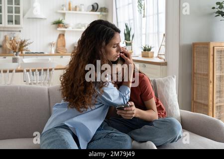 Cura gentile donna hugging sconvolto piangendo ragazza teen siede sul divano con telefono e soffre di stress Foto Stock