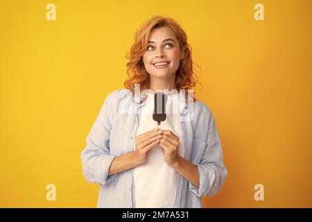 La giovane donna mangia gelati con glassa al cioccolato su sfondo giallo. Divertente donna rossa con gelato. Foto Stock