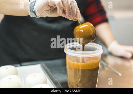 Primo piano di un panettiere con guanti trasparenti che immergono una torta in glassa al caramello. Torte non finite poste su una teglia accanto a una tazza con glassa al caramello. Foto di alta qualità Foto Stock