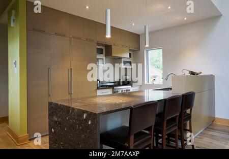 Isola con ripiano in granito grigio scuro e sedie in pelle e legno con schienale alto in cucina all'interno di una casa moderna in stile cubista. Foto Stock