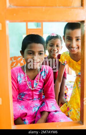 Tre allegre ragazze del Bangladesh, vestite con un vivace abbigliamento tradizionale, sbirciano attraverso un'apertura incorniciata d'arancia, sorridendo brillantemente. Foto Stock
