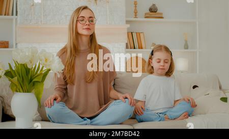 donna caucasica mamma con bambina ragazza seduta a casa sul divano in posizione lotus chiuso occhi meditare insieme. Famiglia meditando madre insegnare Foto Stock