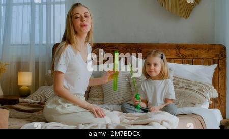 Madre caucasica mamma donna con bambino piccolo bambino ragazza bambino bambino bambino adozione sana figlia soffiando bolle di sapone in aria in camera da letto seduta sul letto Foto Stock